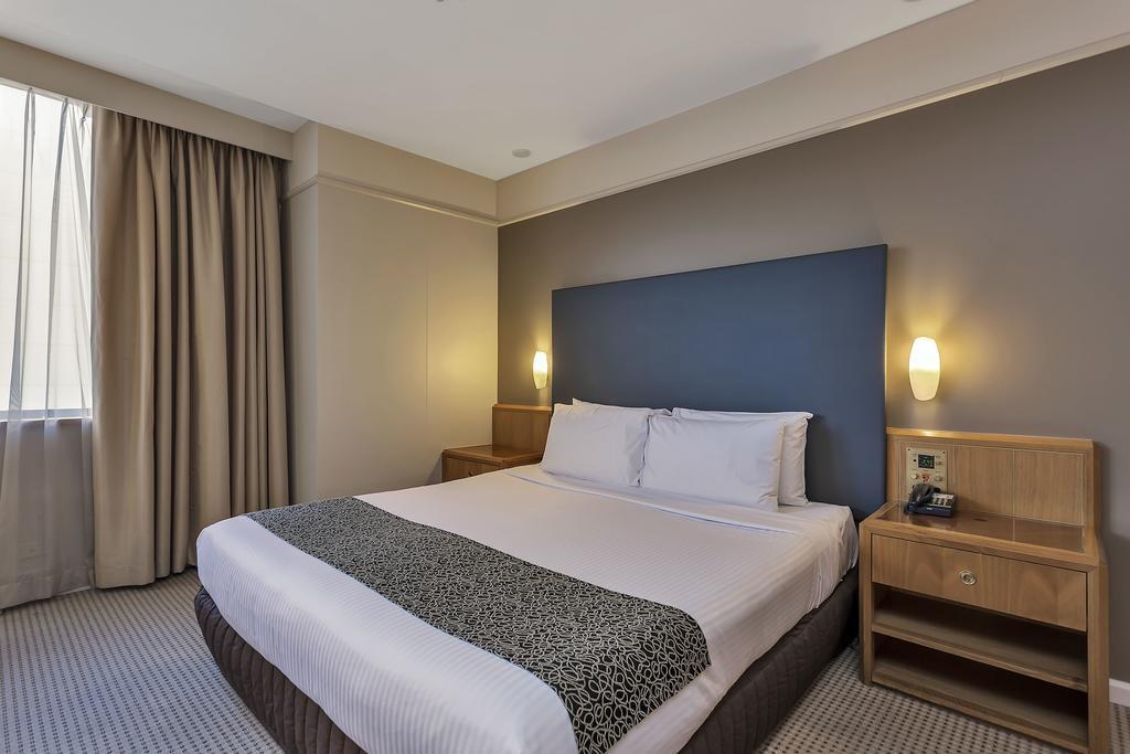 Stamford Plaza Adelaide Hotel ภายนอก รูปภาพ The photo shows a modern hotel room. It features a double bed with a white duvet and a decorative grey and black throw. The headboard is a solid dark color. There are two bedside tables on either side of the bed, each with a lamp. Natural light enter